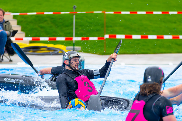 Ein Kanupolo Spieler auf dem Wasser in einem Kanuboot, mit pinker Schwimmweste, schwarzem Helm und schwarzem Paddel. In der rechten Hand hält er den Ball, welcher halb im Wasser liegt
