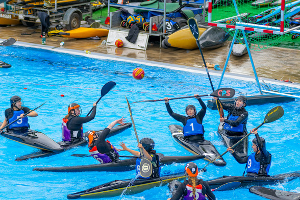 Kanupolo Spielerinnen auf dem Wasser vor dem Tor, welches zwei Meter in der Höhe schwimmt. Eine Spielerin wirft gerade den Ball aufs Tor, eine Spielerin hält ihr Paddel hoch um den ball abzuwehren