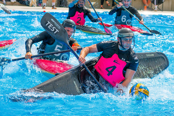 Ein Kanupolo Spieler wird von einem anderen Spieler aus dem Hintergrund mit seinem Boot geschubst; der Vordere hängt fast im Wasser, vor ihm liegt der Ball im Wasser