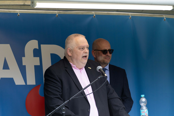Ein dickerer Mann, AfD Northeim Vorstandsvorsitzender Maik Schmitz auf der Bühne vor einem Mikrofon, im Hintergrund ein weiterer Redner und ein blaues Banner mit dem AfD Logo
