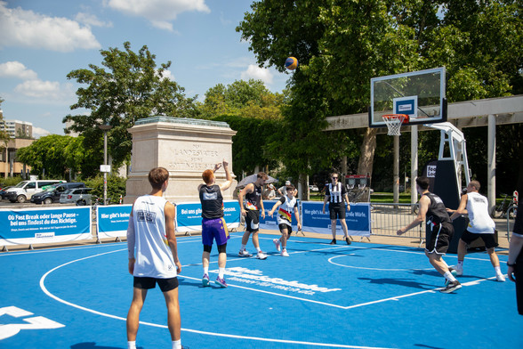 Ein halbes Basketball Feld auf dem Bahnhofsvorplat. Der Boden sind blaue Matten, auf den Matten sechs Spieler, die bei einem Freiwurf grade anlaufen. Im Hintergrund ein Sockel einer Statur und Bäume