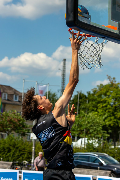 Ein Basketball Spieler mit schwarzen Trikot springt grade zum Korb. Dabei ist er von links hinten zu sehen. Im Hintergrund Baüme und das Bahnhofsgebäude