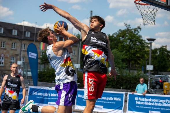 Ein Basketballspieler im weißen Trikot springt zum Korb hoch; ein Spieler in schwarzen Trikot blockt ihm mit einem langen Arm. 
