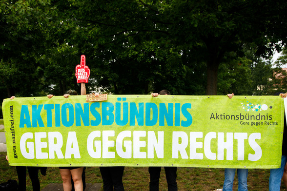 Ein großes Banner wird von Menschen vor grünem Bäumen gehalten; ein großer Flausch Mittelfinder mit der Aufschrift 