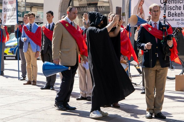 Ein Demonstrant in Mönchskutte protestiert gegen eine Gruppe von Demonstranten vor der Münchner Frauenkirche. Die Demonstranten tragen verschiedene Musikinstrumente, Rosenkränze und rote Schärpen. 