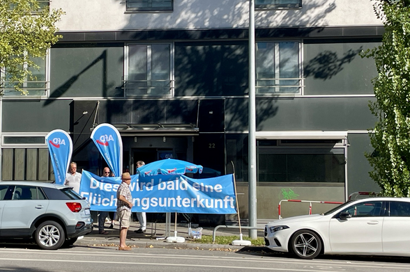 Vor einem ehemaligen Hotel stehen Beach Flags mit AfD Logo, ein blaues leicht zerknittertes Banner mit der Aufschrift Dies wird bald eine Flüchtlingsunterkunft und ein Pavillon. 